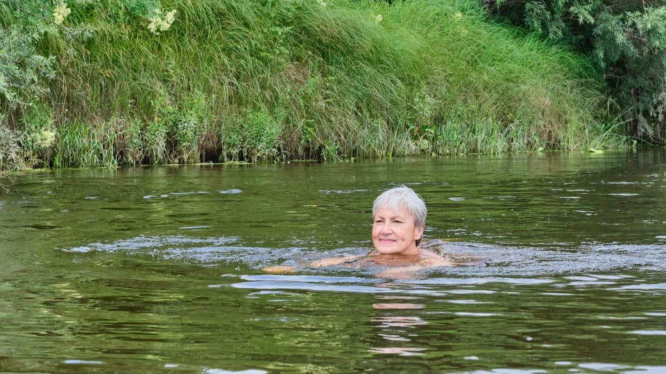 Woman with short grey hair swimming in an open-water lake, surrounded by grassy green bank in the background